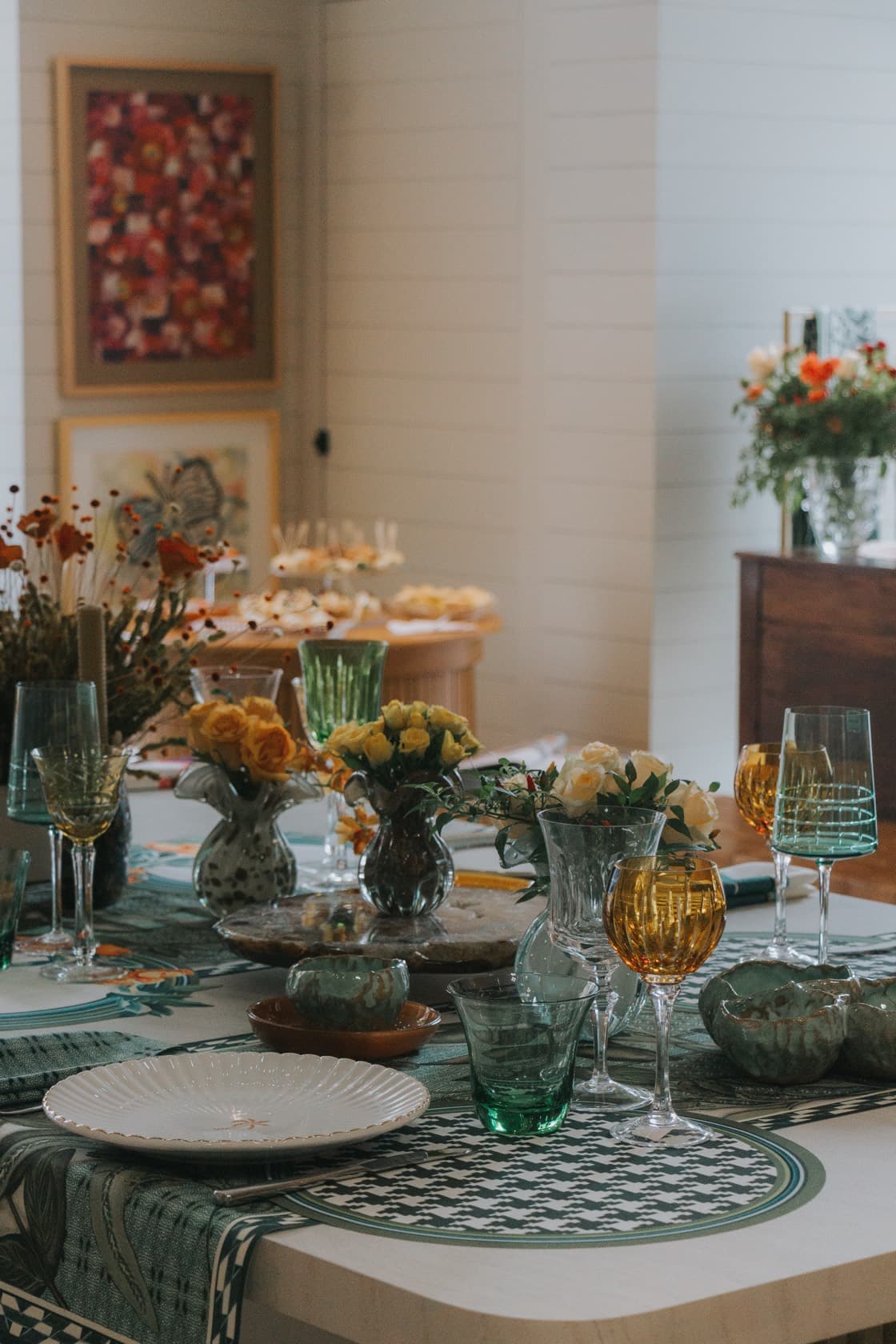 Mesa posta por Carina Verdinelli com taças de cristal verde e âmbar, flores e louça sobre toalha estampada